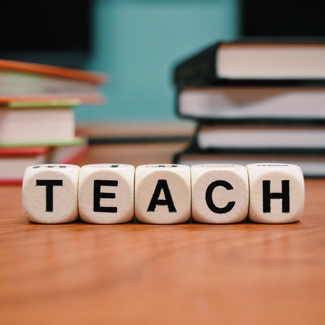 the word teach on wooden dice on a desk with books in the background